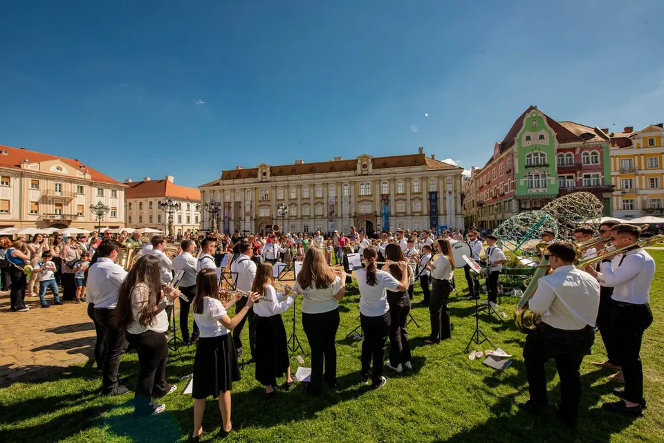 Un grup de muzicieni în haine albe și negre stă pe iarbă, ținând instrumente muzicale în fața unor clădiri colorate sub un cer senin.