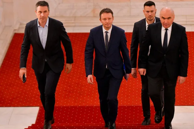 Patru bărbați în costume închise de culoare merg pe un covor roșu în interiorul unei clădiri cu podea de marmură deschisă.