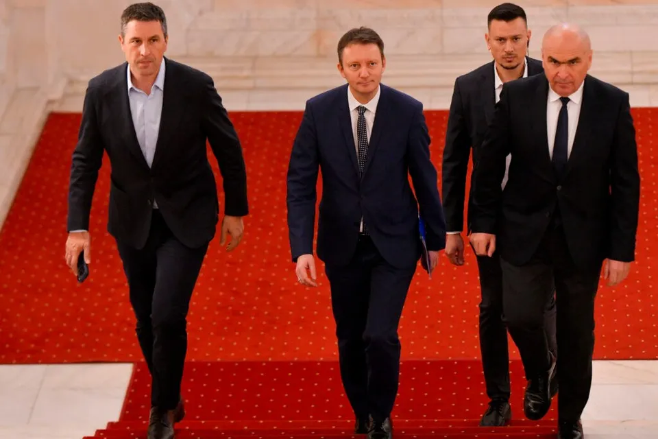 Patru bărbați în costume închise pășesc pe un covor roșu în interiorul unei clădiri.