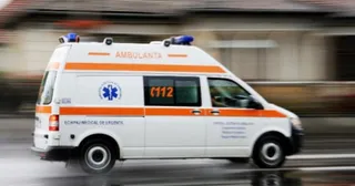 O ambulanță albă cu dungi portocalii și inscripții circulă pe un drum într-o zonă urbană.