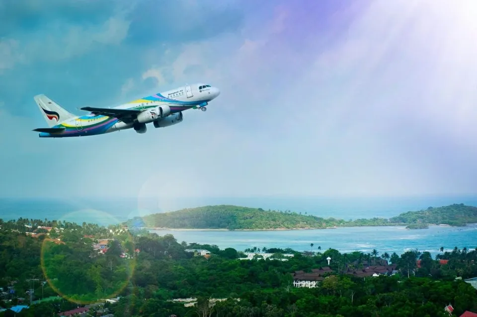 Un avion de pasageri zboară pe cerul senin deasupra unui peisaj de coastă cu vegetație verde și apă albastră.