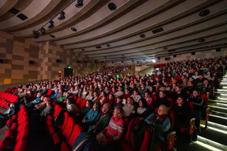 O mulțime de oameni stă așezată în scaune roșii într-o sală de cinema sau teatru întunecată.