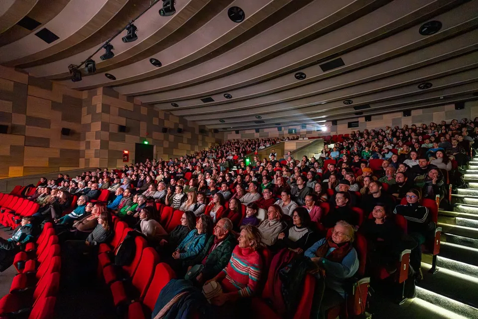 O mulțime de oameni stă așezată în scaune roșii într-o sală de cinema sau teatru întunecată.