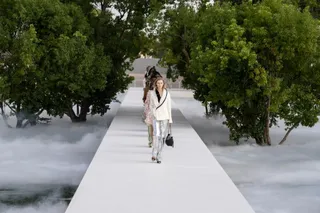 O femeie merge pe un podium alb, înconjurată de copaci verzi și ceață albă, purtând o jachetă albă și pantaloni argintii.
