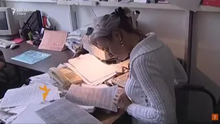 O femeie cu părul prins într-un coc și purtând un pulover alb, stă la un birou plin de hârtii și documente.