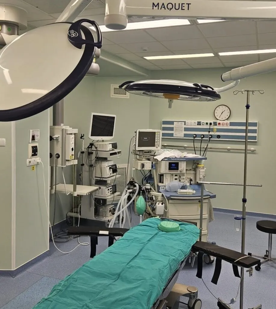 O cameră de operație conține un scaun de examinare acoperit cu un drapel verde, echipamente medicale și lămpi chirurgicale suspendate de tavan.