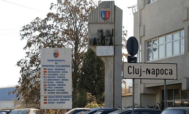 Un semn de stradă indică Cluj-Napoca, lângă un stâlp de beton cu blazon și un panou alb cu o listă de orașe înfrățite.