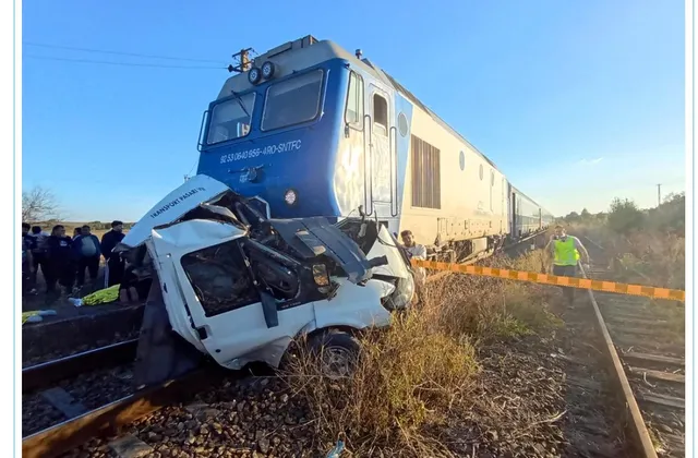 O locomotivă albastră este oprită pe șine lângă o autoutilitară albă zdrobită situată pe o cale ferată cu vegetație uscată în jur.