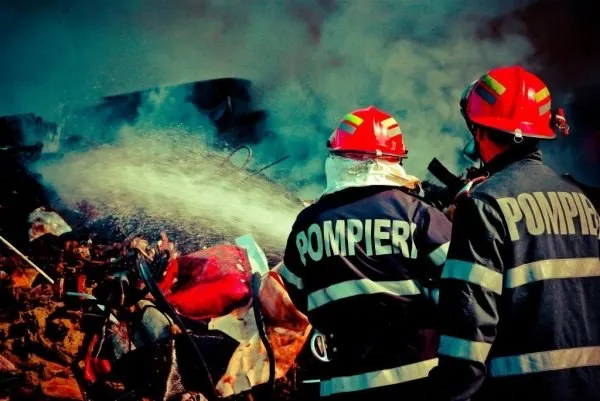 Doi pompieri cu echipament de protecție și căști roșii stau cu spatele și observă un jet de apă care stinge un incendiu cu fum dens.