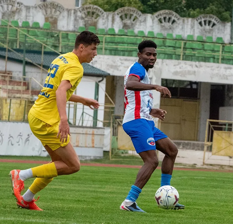 Un jucător în echipament galben aleargă pe terenul de fotbal lângă un jucător în echipament alb și albastru care conduce mingea.