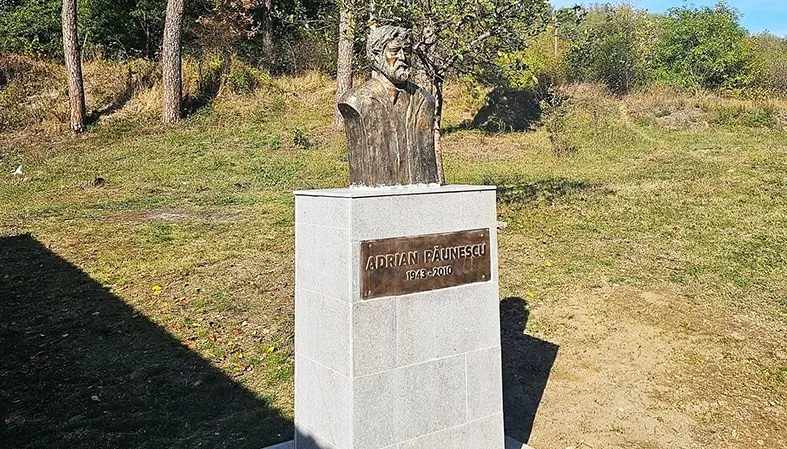 Un bust de bronz cu un bărbat cu barbă este așezat pe un soclu de piatră cu o plăcuță metalică pe un teren cu iarbă.