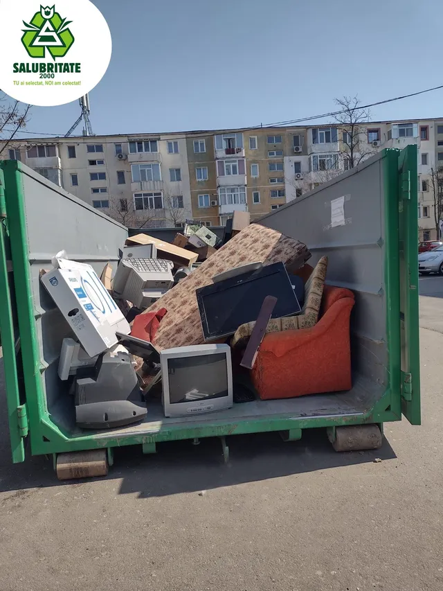 Un container verde conține mobilier uzat și aparate electronice, în fața unor blocuri.