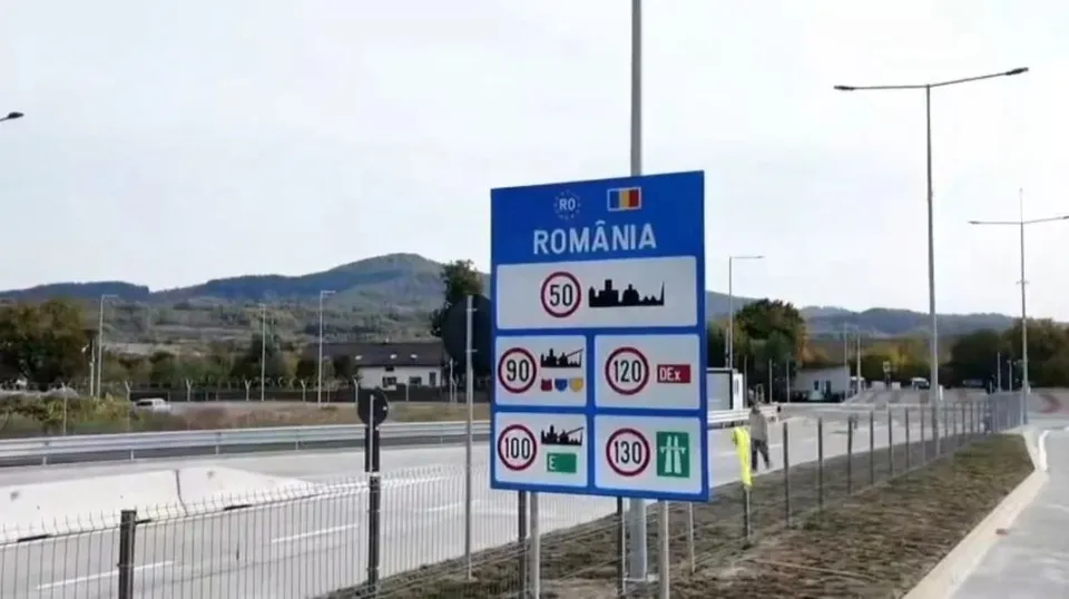 Un panou albastru cu semne de circulație și inscripția ROMÂNIA este amplasat lângă o autostradă cu garduri metalice și dealuri în fundal.