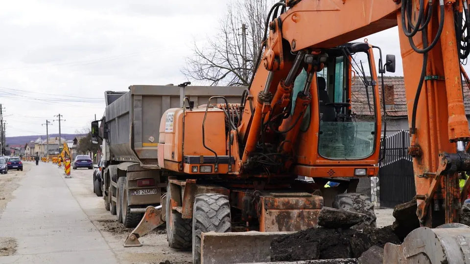 Un excavator portocaliu și un camion basculant sunt parcate pe marginea unui drum, sugerând lucrări de infrastructură. Pe fundal, se văd alte vehicule și clădiri.