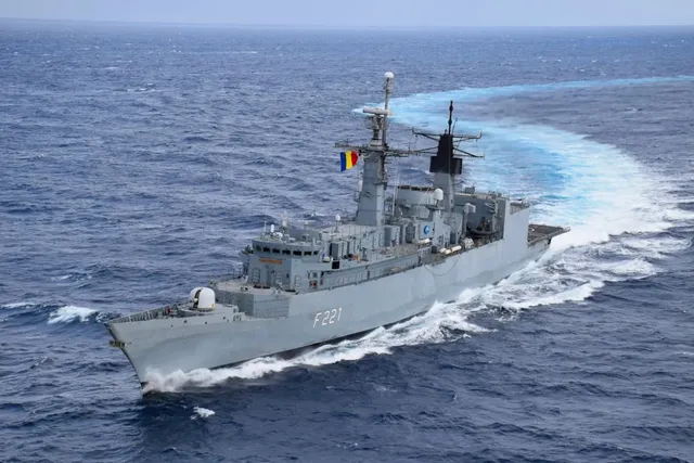 O fregată gri cu numărul F221 navighează pe un ocean albastru, lăsând în urmă un urme de spumă albă.