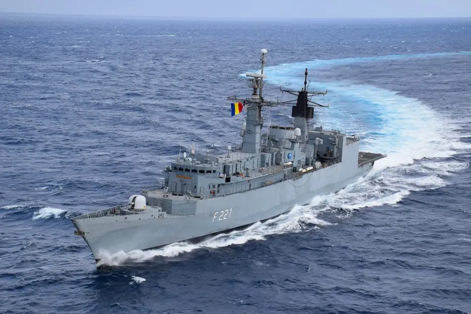 O fregată gri cu numărul F221 navighează pe un ocean albastru, lăsând în urmă un urme de spumă albă.