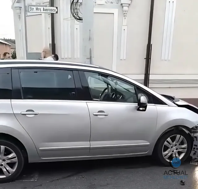 O mașină de culoare argintie este parcată pe lângă un trotuar, în fața unei clădiri albe cu un semn de stradă pe perete.