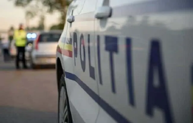 Un vehicul alb cu inscripția POLITIA scrisă pe lateral este parcat pe un drum, având un vehicul argintiu și un om în vestă reflectorizantă în fundal.
