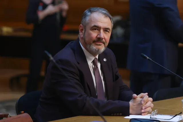 Un bărbat în costum închis, cu cravată și barbă, stă la o masă de lemn cu microfoane, cu brațele încrucișate pe masă.