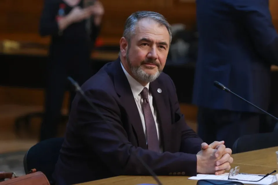 Un bărbat în costum închis, cu cravată și barbă, stă la o masă de lemn cu microfoane, cu brațele încrucișate pe masă.