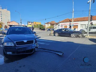 A black Skoda with front-end damage sits in the foreground with debris scattered on the asphalt, while a damaged dark Audi is visible at the intersection in the background under a clear blue sky.