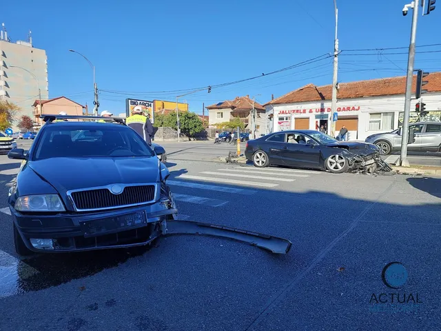 A black Skoda with front-end damage sits in the foreground with debris scattered on the asphalt, while a damaged dark Audi is visible at the intersection in the background under a clear blue sky.