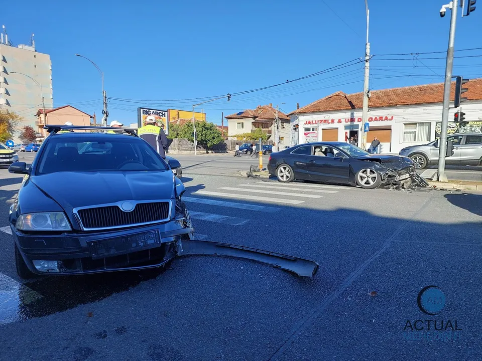 A black Skoda with front-end damage sits in the foreground with debris scattered on the asphalt, while a damaged dark Audi is visible at the intersection in the background under a clear blue sky.