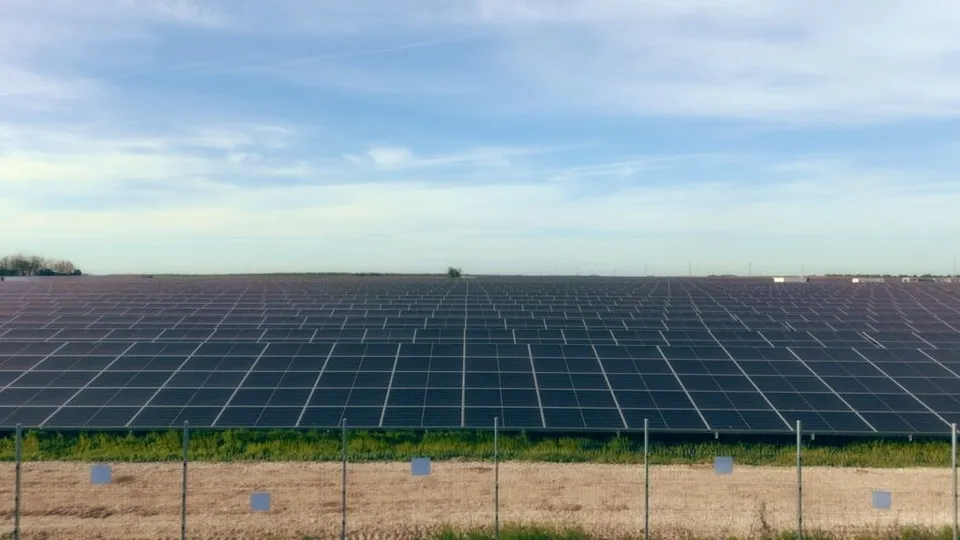 O întinsă de panouri solare negre este instalată pe un teren plat sub un cer cu nori subțiri, fiind delimitată de un gard metalic în prim-plan.