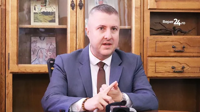 Un bărbat cu părul scurt, purtând sacou albastru, cămașă albă și cravată maro, stă la o masă în fața unui dulap de lemn cu obiecte în vitrină.