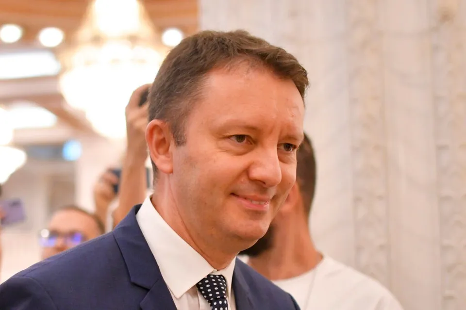 Un bărbat cu părul scurt, purtând sacou albastru, cămașă albă și cravată cu puncte albe, zâmbește într-un decor interior iluminat.