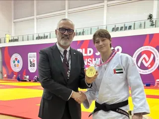 Un bărbat într-un costum negru strânge mâna unei femei îmbrăcate într-un giu de judo alb care poartă o medalie aurie și un cordon.