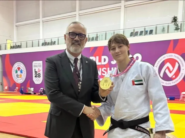 Un bărbat într-un costum negru strânge mâna unei femei îmbrăcate într-un giu de judo alb care poartă o medalie aurie și un cordon.