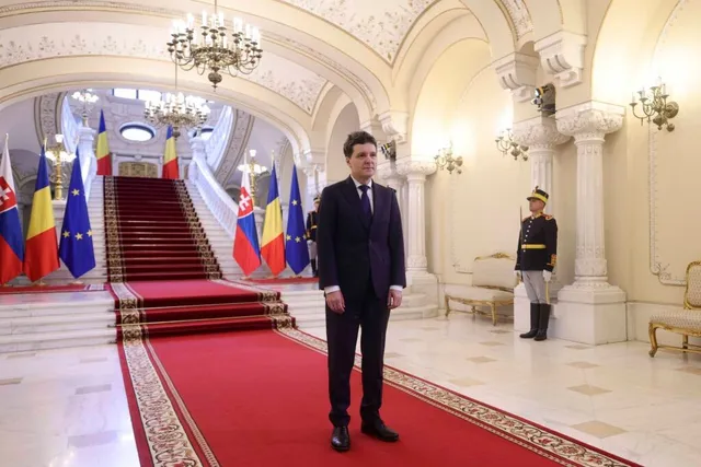 Un bărbat într-un costum închis la culoare stă pe un covor roșu într-o încăpere cu coloane, scări late și candelabre suspendate.