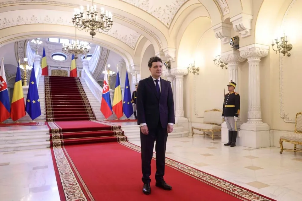 Un bărbat într-un costum închis la culoare stă pe un covor roșu într-o încăpere cu coloane, scări late și candelabre suspendate.
