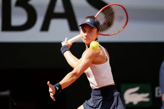 O femeie cu o șapcă neagră și o cană albă ține o raketă de tenis în timp ce o minge galbenă zboară în fața ei.