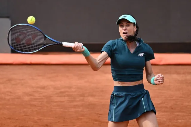 O femeie cu șapcă și echipament sportiv de culoare închisă lovește o minge galbenă cu o rachetă pe un teren de culoarea cărămizii.