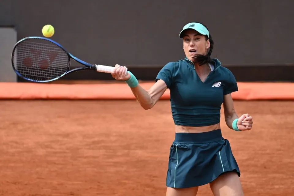 O femeie cu șapcă și echipament sportiv de culoare închisă lovește o minge galbenă cu o rachetă pe un teren de culoarea cărămizii.