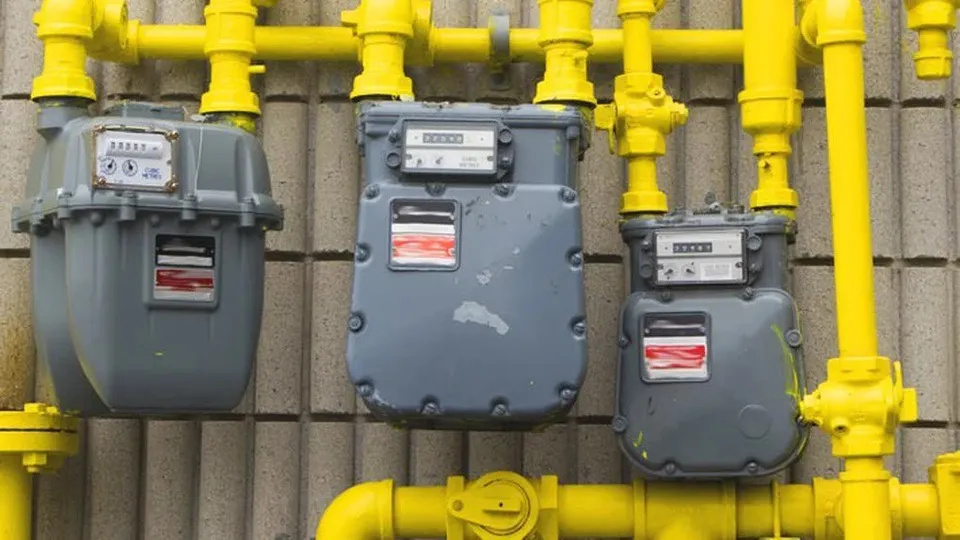 Trei contoare de gaz gri sunt montate pe o rețea de țevi galbene dispuse orizontal și vertical pe un perete de beton.