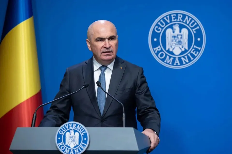 Un bărbat cu părul scurt și deschis la tâmple poartă costum închis la culoare, cămașă albă și cravată albastră în fața unui fundal albastru cu un emblemă circulară.
