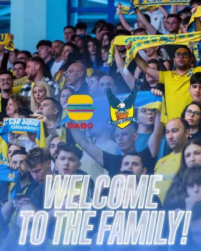 O mulțime de oameni stă în tribune, unii purtând tricouri galbene și ridicând steaguri colorate sub un text alb mare.