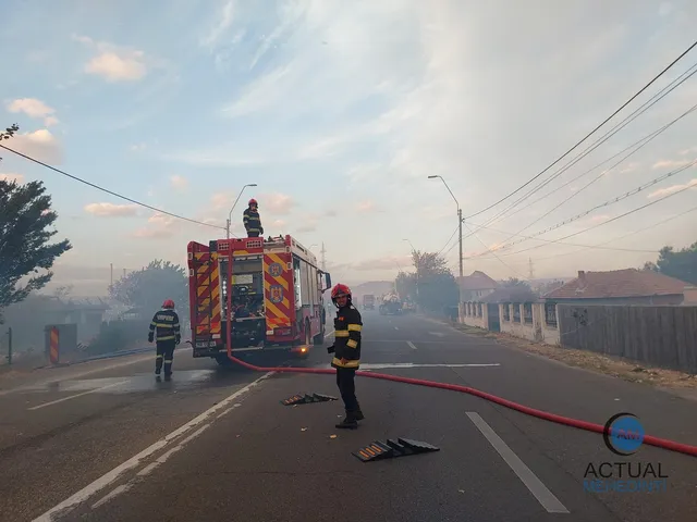 Pompieri în echipament de protecție stau lângă un autospecială de stingere parcată pe un drum asfaltat sub un cer înnorat.