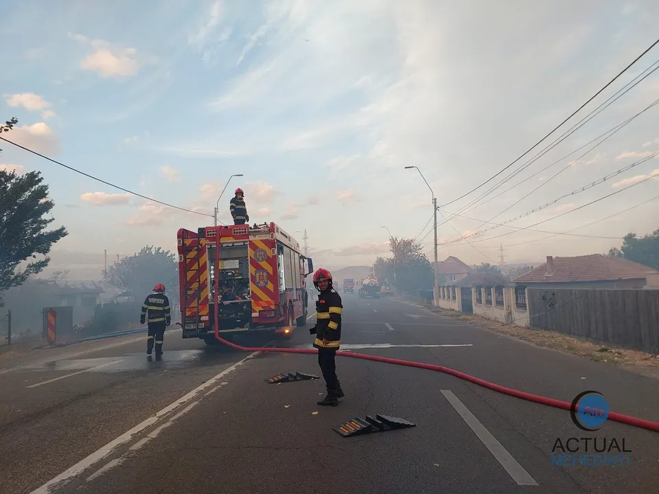 Pompieri în echipament de protecție stau lângă un autospecială de stingere parcată pe un drum asfaltat sub un cer înnorat.