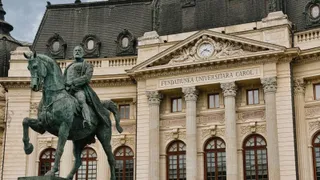 O statuie ecuestre de culoare închisă este amplasată în fața unei clădiri monumentale cu coloane și inscripție pe fațadă.