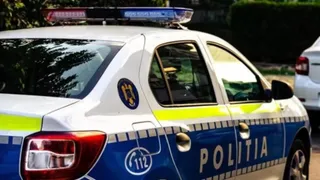 Un autoturism alb cu marcaje albastre și galbene, pe care scrie POLITIA, este parcat pe un drum.