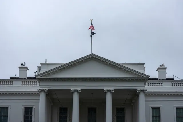 Imaginea arată fațada Casei Albe, cu acoperiș, fronton, coloane, ferestre și steag american vizibil pe cerul înnorat.