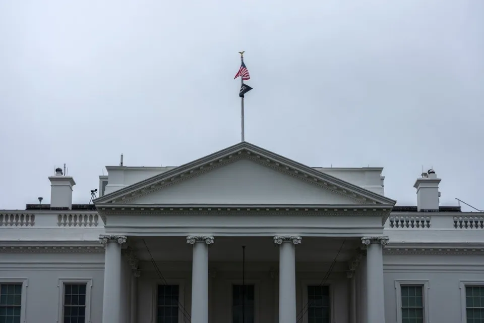 Imaginea arată fațada Casei Albe, cu acoperiș, fronton, coloane, ferestre și steag american vizibil pe cerul înnorat.