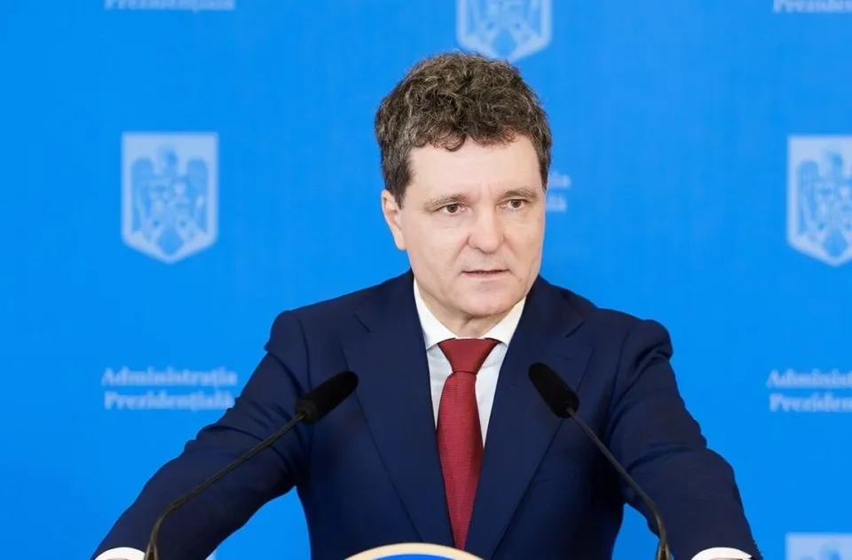 Un bărbat cu părul creț, purtând un costum albastru și o cravată roșie, stă în fața unui fundal albastru cu embleme albe.