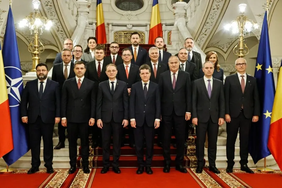 Un grup de bărbați în costumuri de formalitate, în picioare pe un covor roșu și scări, într-o sală mare cu steaguri, un candelabru și o balustradă de marmură.