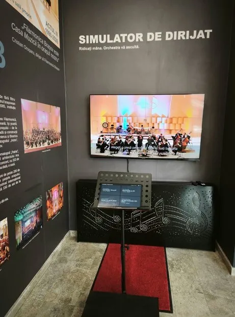 Un ecran digital afișează o orchestră pe un perete negru, lângă un suport de partitură și un panou cu note muzicale.