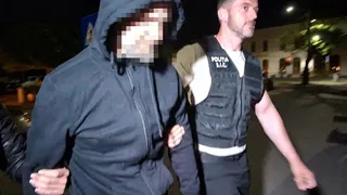 Un bărbat cu vestă neagră marcată cu text alb conduce un individ cu capotă și haine închise într-un mediu exterior noaptea.
